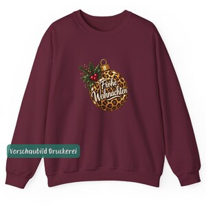 May include: A burgundy long-sleeve sweatshirt featuring a central graphic of a round Christmas ornament with a brown and black leopard print pattern. The ornament is adorned with green holly leaves and red berries, and displays the text Frohe Weihnachten.