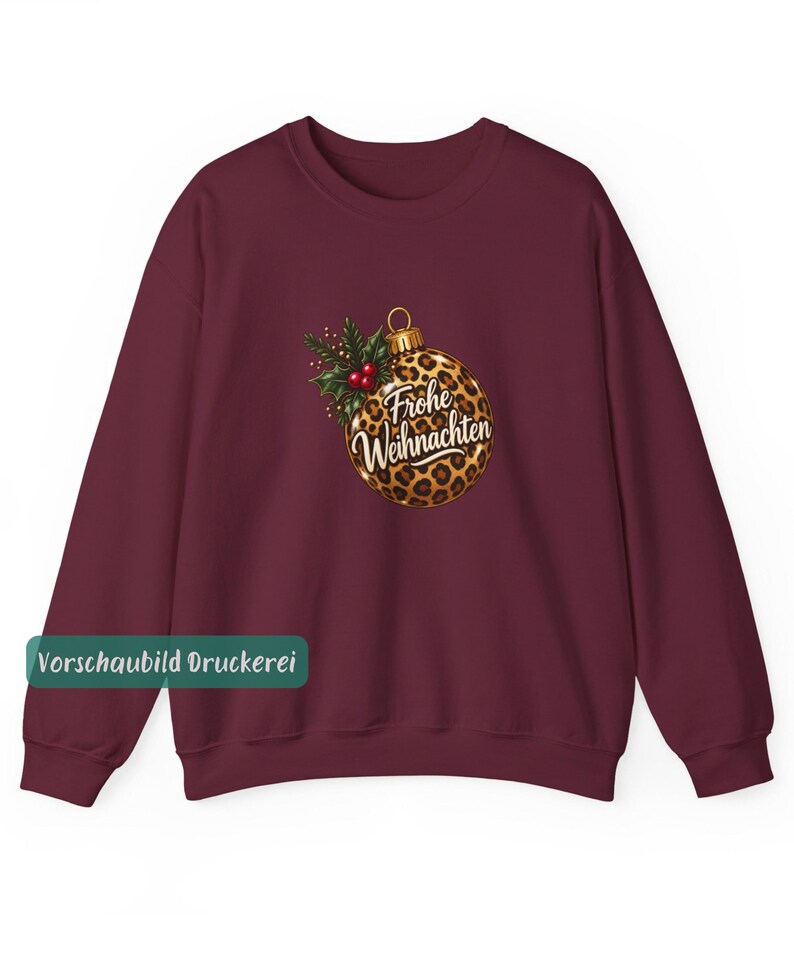 May include: A burgundy long-sleeve sweatshirt featuring a central graphic of a round Christmas ornament with a brown and black leopard print pattern. The ornament is adorned with green holly leaves and red berries, and displays the text Frohe Weihnachten.