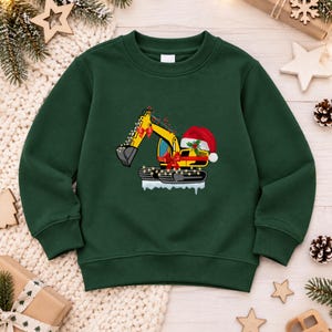 May include: A dark green long-sleeve sweatshirt featuring a graphic of a yellow excavator decorated with Christmas lights, red bows, and a Santa hat. The excavator sits on a patch of white snow, creating a festive holiday-themed design.
