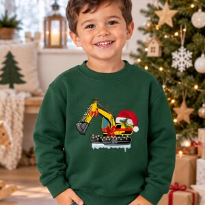 May include: A dark green crewneck sweatshirt featuring a graphic of a yellow excavator decorated with red bows, string lights, and a red Santa hat, positioned on a patch of white snow, worn with blue denim jeans.