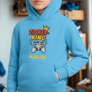 May include: A light blue hooded sweatshirt featuring a graphic print with a yellow crown, the text ZOCKER KING GAME ON, a grey video game controller illustration, and the name MARLON printed in yellow below the controller graphic.