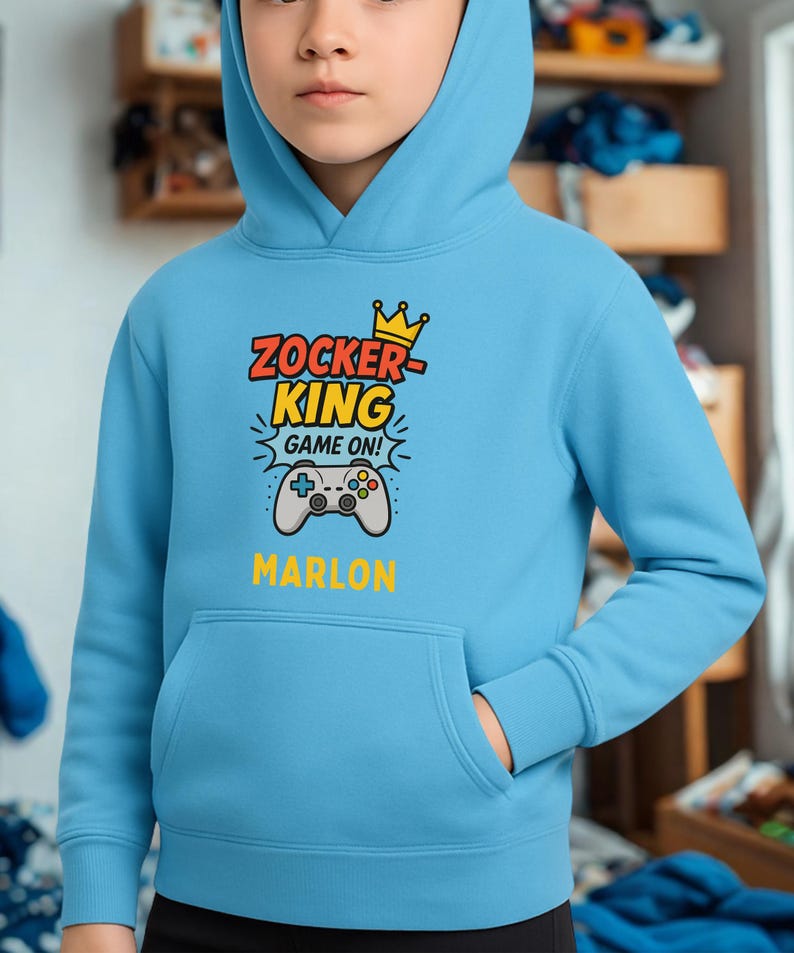 May include: A light blue hooded sweatshirt featuring a graphic print with a yellow crown, the text ZOCKER KING GAME ON, a grey video game controller illustration, and the name MARLON printed in yellow below the controller graphic.