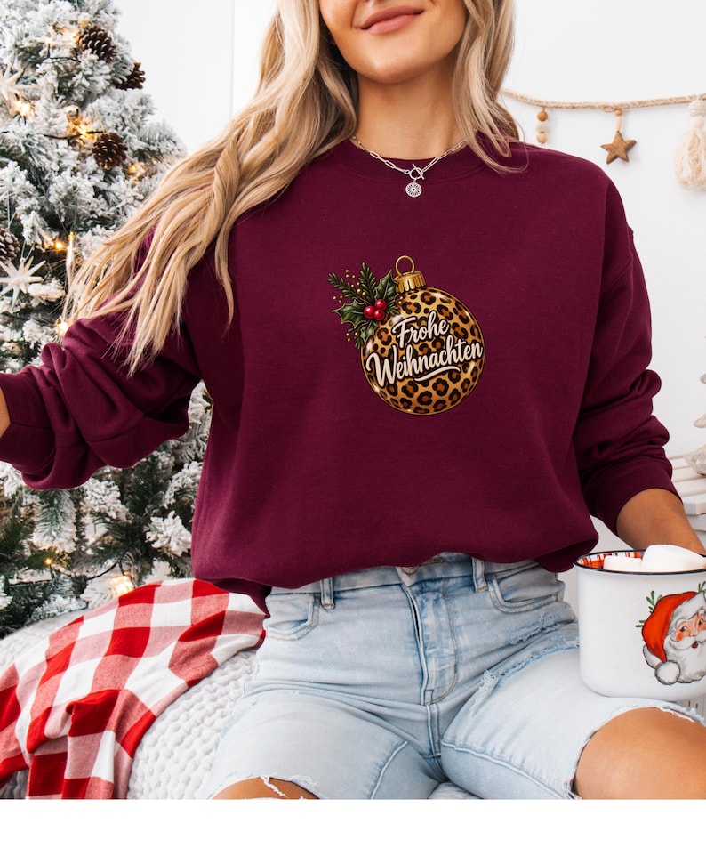 May include: A maroon long-sleeve sweatshirt featuring a central graphic of a leopard print Christmas ornament. The ornament is adorned with green holly leaves and red berries, and includes the white cursive text Frohe Weihnachten.
