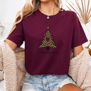 K&ouml;nnte beinhalten: Ein bordeauxrotes Kurzarm-T-Shirt mit einer Grafik eines Weihnachtsbaums mit Leopardenmuster, einem kleinen herzf&ouml;rmigen Ausschnitt und einem Stern an der Spitze. Der Text "merry christmas" ist in schwarzer Schreibschrift unter dem Baum.