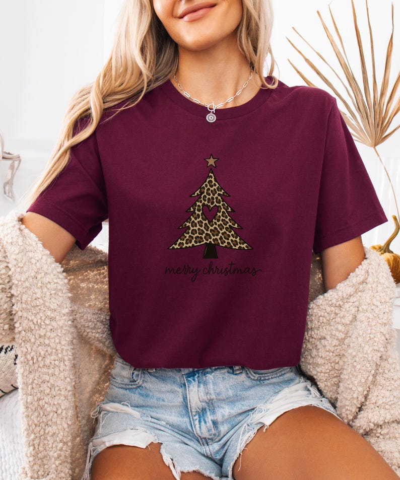 K&ouml;nnte beinhalten: Ein bordeauxrotes Kurzarm-T-Shirt mit einer Grafik eines Weihnachtsbaums mit Leopardenmuster, einem kleinen herzf&ouml;rmigen Ausschnitt und einem Stern an der Spitze. Der Text "merry christmas" ist in schwarzer Schreibschrift unter dem Baum.