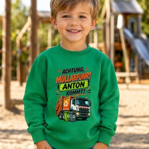 May include: A bright green long-sleeve sweatshirt featuring a graphic print of an orange and white garbage truck with two small trash bins. The text reads ACHTUNG, M&Uuml;LLABFUHR! ANTON KOMMT! in orange, black, and white lettering on the front.