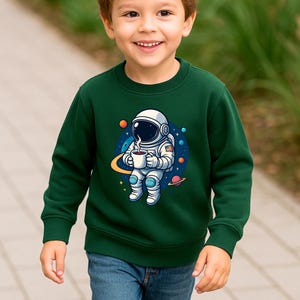 May include: A dark green long-sleeve sweatshirt featuring a graphic print of an astronaut in a white suit holding a steaming mug of coffee, surrounded by colorful planets, stars, and a yellow orbital ring against a dark blue circular background.