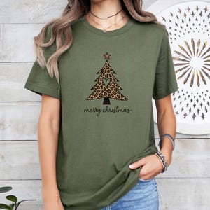 K&ouml;nnte beinhalten: Ein olivgr&uuml;nes T-Shirt mit einer Grafik eines Weihnachtsbaums mit Leopardenmuster, einem kleinen herzf&ouml;rmigen Ausschnitt in der Mitte und einem Stern an der Spitze. Der Text merry christmas ist in schwarzer Schreibschrift darunter gedruckt.