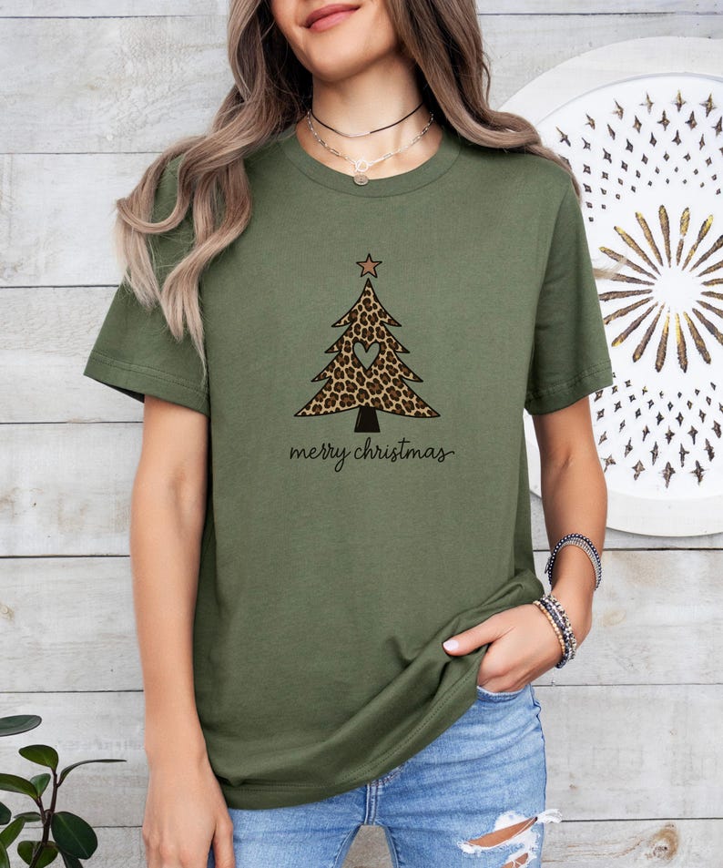 K&ouml;nnte beinhalten: Ein olivgr&uuml;nes T-Shirt mit einer Grafik eines Weihnachtsbaums mit Leopardenmuster, einem kleinen herzf&ouml;rmigen Ausschnitt in der Mitte und einem Stern an der Spitze. Der Text merry christmas ist in schwarzer Schreibschrift darunter gedruckt.