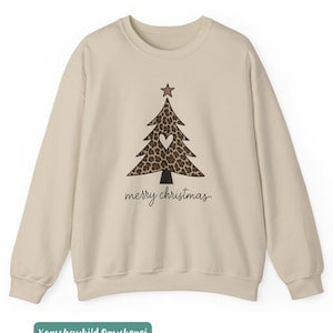 May include: A beige crewneck sweatshirt featuring a graphic of a Christmas tree with a brown leopard print pattern, a white heart in the center, and a brown star on top. The text merry christmas is printed in black cursive script below the tree.