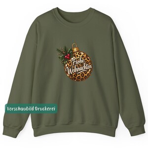 May include: An olive green crewneck sweatshirt featuring a central graphic of a round Christmas ornament with a leopard print pattern. The ornament is decorated with green holly leaves and red berries, and displays the text Frohe Weihnachten.