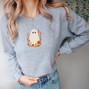 May include: A heather gray crewneck sweatshirt featuring a central graphic of a cream-colored ghost surrounded by orange and brown autumn-themed flowers, leaves, and wheat stalks. The long-sleeved garment has a ribbed neckline, cuffs, and hem.