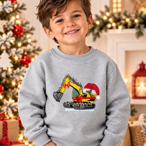 May include: A heather gray crewneck sweatshirt featuring a graphic of a yellow excavator decorated with red bows, green pine branches, and a red Santa hat. The excavator tracks are wrapped in small string lights, and the design sits on a white base.