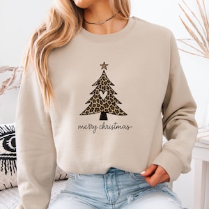 May include: A beige crewneck sweatshirt featuring a graphic of a Christmas tree with a leopard print pattern, a small white heart in the center, and a star on top. Below the tree, the text merry christmas is printed in a black cursive font.