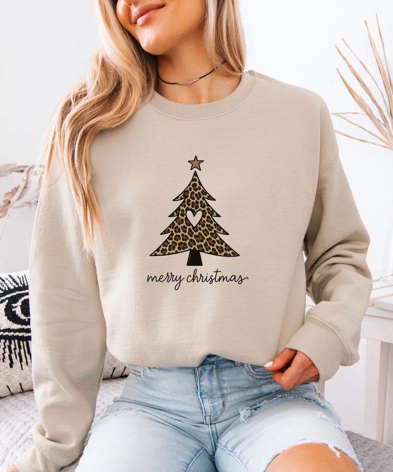 May include: A beige crewneck sweatshirt featuring a graphic of a Christmas tree with a leopard print pattern, a small white heart in the center, and a star on top. Below the tree, the text merry christmas is printed in a black cursive font.