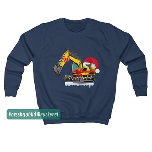May include: A navy blue long-sleeve sweatshirt featuring a graphic of a yellow excavator decorated with red bows, holly, and string lights. The excavator wears a red Santa hat and sits on a patch of white snow. The text "Vorschaubild Druckerei" is visible.
