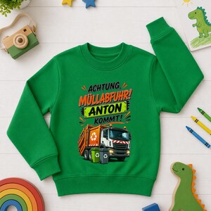 May include: A bright green long-sleeve sweatshirt featuring a graphic of an orange and white garbage truck with two trash bins. The text reads "ACHTUNG, M&Uuml;LLABFUHR! ANTON KOMMT!" in black and orange lettering, with the name "ANTON" on a green background.