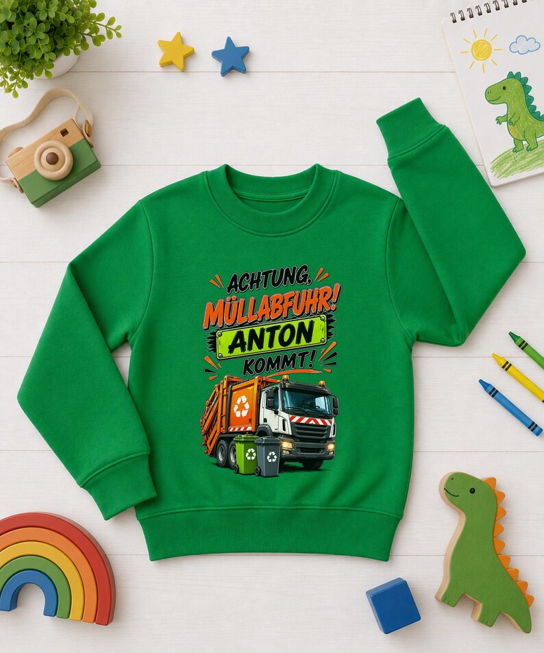 May include: A bright green long-sleeve sweatshirt featuring a graphic of an orange and white garbage truck with two trash bins. The text reads "ACHTUNG, M&Uuml;LLABFUHR! ANTON KOMMT!" in black and orange lettering, with the name "ANTON" on a green background.