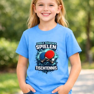 Puede incluir: Una camiseta azul claro con el texto alem&aacute;n "COOLE M&Auml;DCHEN SPIELEN TISCHTENNIS". El gr&aacute;fico incluye una raqueta de ping-pong roja y azul, una pelota blanca y un dise&ntilde;o de salpicaduras azules. La camiseta la lleva una persona joven.