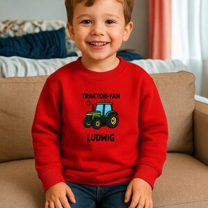May include: A bright red long-sleeved sweatshirt featuring a graphic of a green tractor in the centre. Above the tractor, the text reads TRAKTOR-FAN, and below the tractor, the name LUDWIG is printed in bold black capital letters.