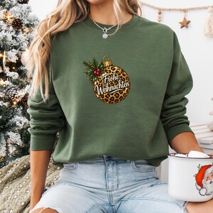 May include: An olive green crewneck sweatshirt featuring a central graphic of a leopard print Christmas ornament. The ornament is adorned with green holly leaves and red berries, with the German text Frohe Weihnachten printed in black script.