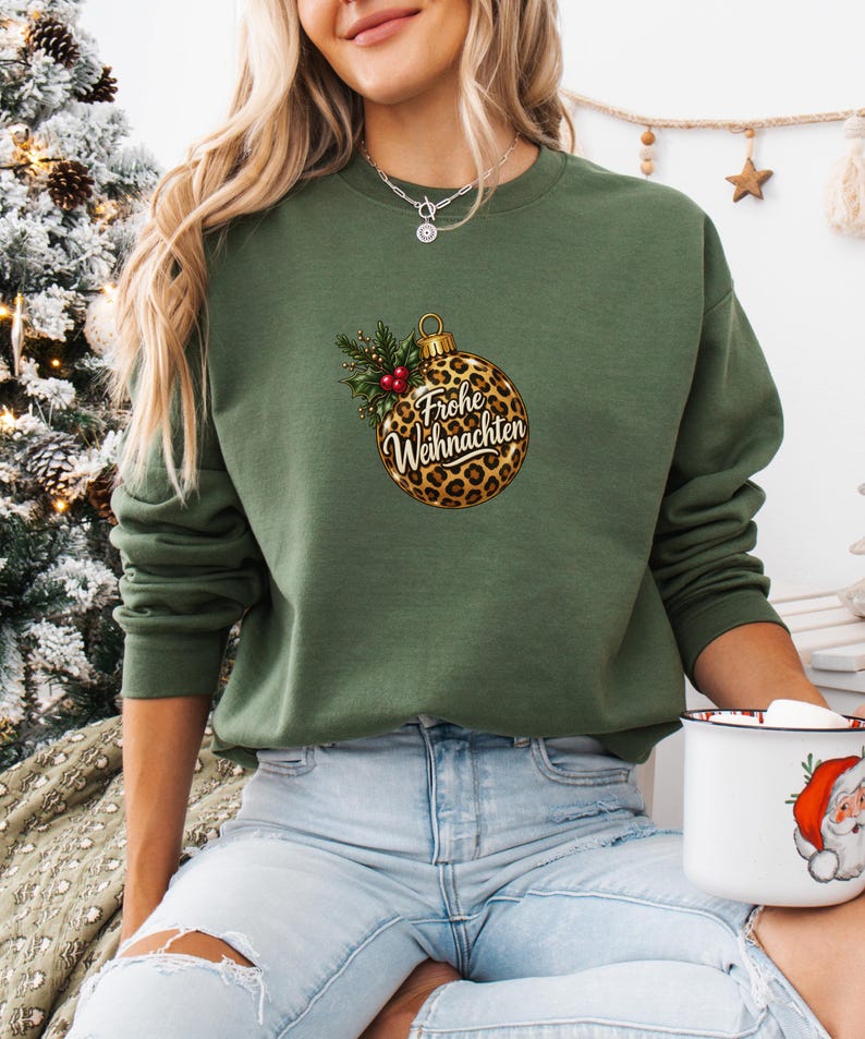 May include: An olive green crewneck sweatshirt featuring a central graphic of a leopard print Christmas ornament. The ornament is adorned with green holly leaves and red berries, with the German text Frohe Weihnachten printed in black script.
