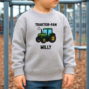 May include: A heather grey long-sleeve sweatshirt featuring a graphic of a green and yellow tractor with blue windows. Above the tractor, the text reads TRAKTOR-FAN, and below the tractor, the name WILLY is printed in bold black capital letters.