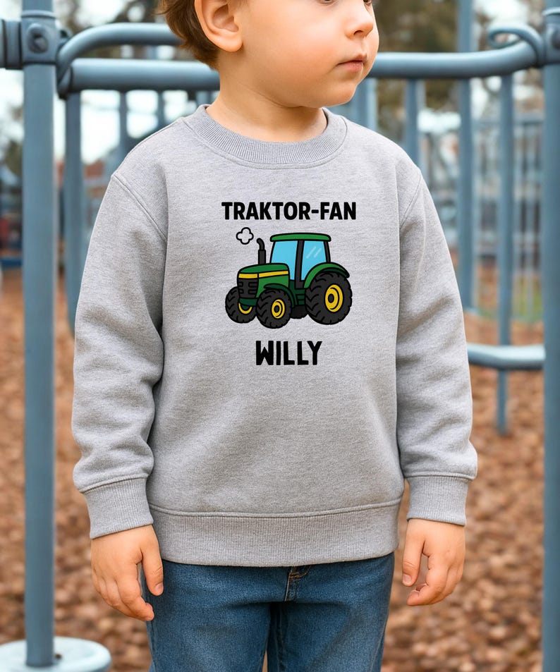 May include: A heather grey long-sleeve sweatshirt featuring a graphic of a green and yellow tractor with blue windows. Above the tractor, the text reads TRAKTOR-FAN, and below the tractor, the name WILLY is printed in bold black capital letters.