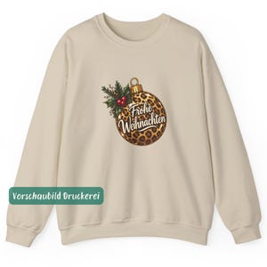 May include: A beige crewneck sweatshirt featuring a central graphic of a Christmas ornament with a leopard print pattern. The ornament is adorned with green holly leaves and red berries, and displays the white cursive text Frohe Weihnachten.