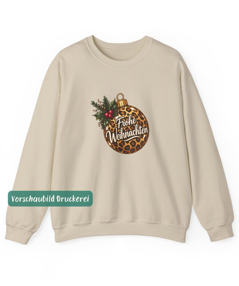 May include: A beige crewneck sweatshirt featuring a central graphic of a Christmas ornament with a leopard print pattern. The ornament is adorned with green holly leaves and red berries, and displays the white cursive text Frohe Weihnachten.
