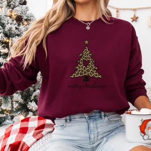 May include: A maroon long-sleeve sweatshirt featuring a graphic of a Christmas tree with a leopard print pattern, a central heart cutout, and a star topper. The text "merry christmas" is printed in black cursive script below the tree design.