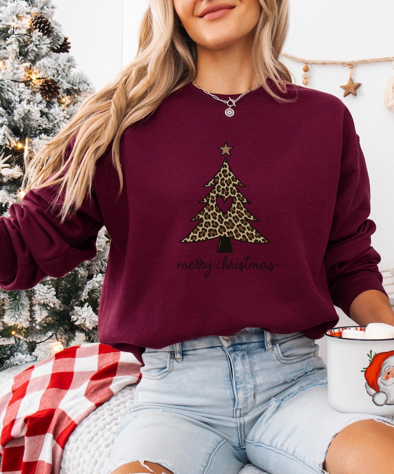 May include: A maroon long-sleeve sweatshirt featuring a graphic of a Christmas tree with a leopard print pattern, a central heart cutout, and a star topper. The text "merry christmas" is printed in black cursive script below the tree design.