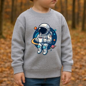 May include: A heather gray long-sleeve sweatshirt featuring a graphic print of an astronaut in a white suit holding a steaming mug. The astronaut is positioned against a dark blue circular background with colorful planets, stars, and a yellow ring.