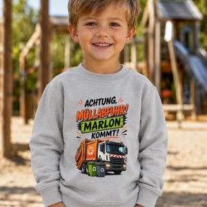 May include: A light gray long-sleeve sweatshirt featuring a graphic of an orange and white garbage truck with two small trash bins. The text reads ACHTUNG, M&Uuml;LLABFUHR! MARLON KOMMT! in orange, black, and green lettering on the front of the garment.