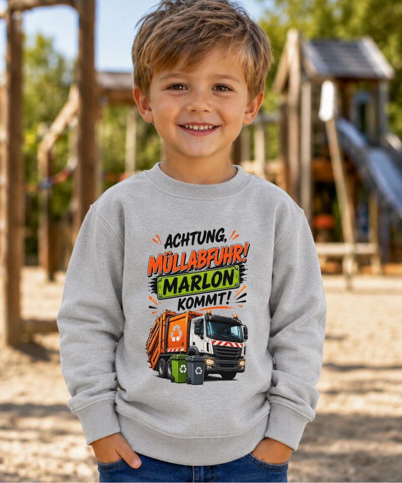 May include: A light gray long-sleeve sweatshirt featuring a graphic of an orange and white garbage truck with two small trash bins. The text reads ACHTUNG, M&Uuml;LLABFUHR! MARLON KOMMT! in orange, black, and green lettering on the front of the garment.