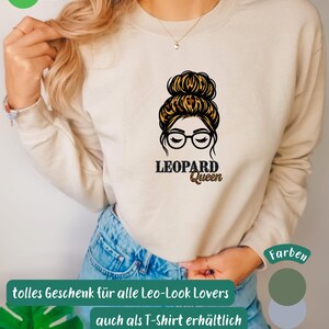 Könnte beinhalten: Ein beiges Langarm-Sweatshirt mit einem Grafikdruck einer Person mit Dutt-Frisur und Brille. Das Haar weist ein Leopardenmuster auf. Unter der Grafik steht der Text LEOPARD Queen in schwarzen Buchstaben und mit Leopardenmuster-Optik.