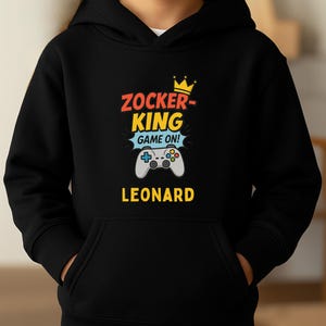 May include: A black hooded sweatshirt featuring a graphic print with the text ZOCKER-KING GAME ON! and LEONARD. The design includes a yellow crown icon above the text and a gray video game controller illustration centered below the text.