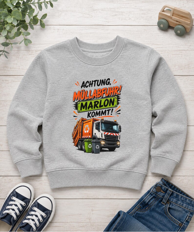 May include: A heather gray long-sleeve sweatshirt featuring a graphic of an orange garbage truck with two recycling bins. The text reads "ACHTUNG, M&Uuml;LLABFUHR! MARLON KOMMT!" with "MARLON" displayed on a green rectangular sign above the truck.