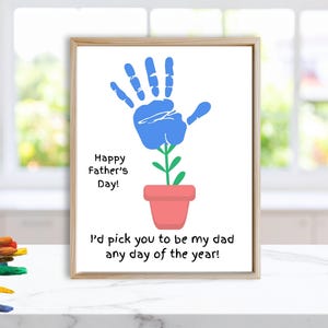 Può includere: Opera d'arte incorniciata con un disegno di impronta di mano blu, che ricorda un fiore. Il testo "Happy Father's Day!" e "I'd pick you to be my dad any day of the year!" sono inclusi. La cornice è marrone chiaro.
