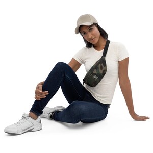 Könnte beinhalten: Eine Person trägt ein weißes T-Shirt, blaue Jeans und eine beige Baseballkappe und sitzt auf weißem Hintergrund. Sie trägt eine Camouflage-Bauchtasche mit einem schwarzen Gurt.