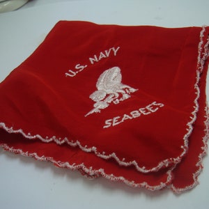 May include: A folded red fabric square with a scalloped white stitched edge. The center features white embroidery depicting the U.S. Navy Seabees emblem, with the text U.S. NAVY arched above and the word SEABEES embroidered below the central graphic.