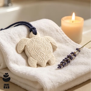 May include: A white ceramic turtle ornament featuring intricate embossed spiral and geometric patterns. The turtle is suspended by a dark blue cord and rests on a folded white towel next to a sprig of dried lavender and a lit candle in a glass.