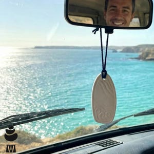 May include: Cream oval car air freshener with a wave pattern, suspended from a rearview mirror. The background features a blue sea and a person smiling in the mirror.