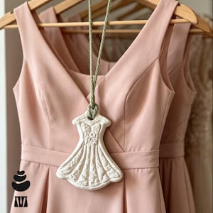 May include: A pale pink sleeveless dress with a V-neckline and a defined waistline, featuring a white ceramic ornament shaped like a dress hanging from a sage green cord. The ceramic piece displays a detailed floral pattern on its surface.