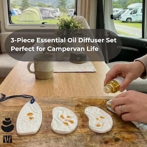 May include: A 3-piece essential oil diffuser set with white ceramic diffusers and a wooden base. The set includes a bottle of oil being poured onto a diffuser. The text reads "3-Piece Essential Oil Diffuser Set Perfect for Campervan Life."