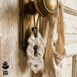 May include: A white, textured, decorative keyhole escutcheon ornament hangs from a dark green cord attached to a tarnished brass door knob and backplate. The ornament features a central keyhole cutout and a smaller circular hole at the bottom.