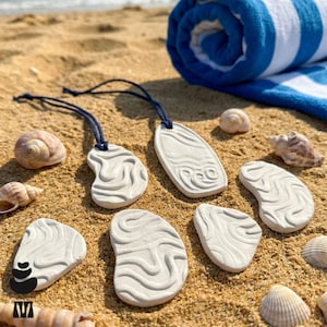 May include: A collection of white clay ornaments with embossed wavy patterns, including two pieces with dark blue hanging cords, arranged alongside various seashells on a sandy surface with a blue and white striped towel in the background.