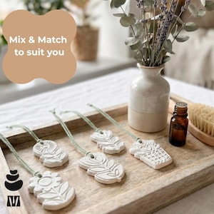 May include: Four white clay ornaments with embossed floral and botanical designs, each attached to a sage green string. The ornaments are arranged on a wooden tray alongside a small ceramic vase, a brown glass essential oil bottle, and a brush.
