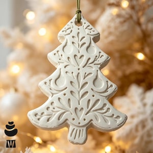 May include: A white, tree-shaped ornament featuring intricate, embossed floral and heart patterns. The ornament has a smooth, matte finish and is suspended by a thin, olive-green cord attached to a small hole at the top of the tree structure.