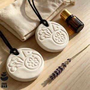 May include: Two circular white clay ornaments featuring embossed floral and geometric patterns, each suspended by a black cord. A small amber glass bottle with a gold cap and a sprig of dried lavender are positioned nearby on a light wood surface.